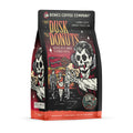 The front of a 12 ounce bag of Bones Coffee Company From Dusk til Dawn coffee. Its flavor is jelly donut, and it has a skeleton dressed like a vampire holding a jelly donut while people leave a donut shop on the art.