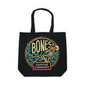A black tote bag that has the classic Bones Coffee Company logo on it. The logo has a skeleton drinking coffee resting on greenery on it.