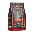 The front of a 12 ounce bag of Bones Coffee Company White Russian coffee. Its flavor is cream and coffee liqueur, and it has a skeleton wearing sunglasses with a goatee in a comfy coat with a white tee shirt holding a cup of coffee on the art.